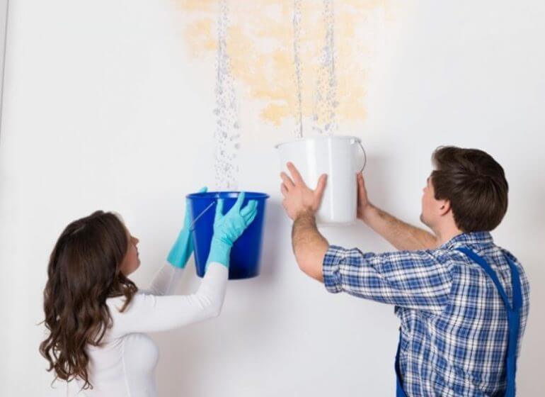 A man and woman try to stop a leaking roof, armed with buckets to catch the falling water.